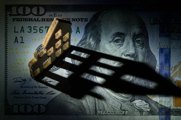 Geleneksel bir ahşap ev 100 Amerikan Doları banknotunun bir parçasının üzerinde duruyor ve fatura üzerinde uzun bir gölge bırakıyor..