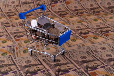 100 dolarlık banknotların arka planında farklı ülkelerin bozuk paraları olan bir süpermarketten alınmış minyatür bir alışveriş arabası.