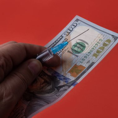 Sıvı dolu plastik tek kullanımlık şırınga ve kırmızı arka planda 100 dolarlık banknot tutan erkek parmakları.