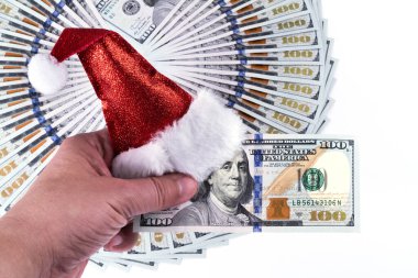 Bir adam 100 Amerikan Doları 'nı elinde tutuyor. Kırmızı minyatür bir noel şapkası ve arka planda daire şeklinde katlanmış 100 dolarlık banknotlar var.