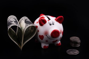 Komik domuz kumbarası, siyah arka planda insan kalbi sembolü şeklinde iki Amerikan dolarının yanında.