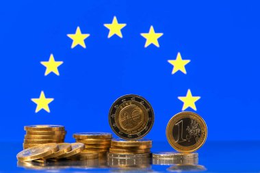 Kenarda duran 1 ve 2 avroluk madeni paralar, Avrupa Birliği bayrağının arka planına karşı çok sayıda diğer madeni para