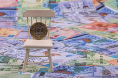 A coin in denomination of 100 Kazakhstani tenge stands on its edge on a miniature chair against the background of Kazakhstani banknotes clipart