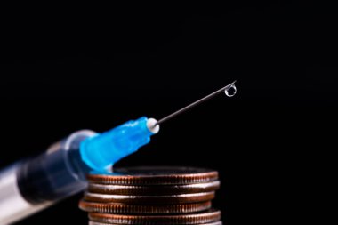 Tek kullanımlık şırınga, iğnelerin ucunda bir damla sıvı ve siyah arka planda bir sütun bozuk para.