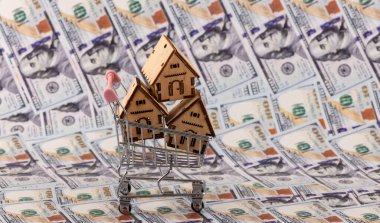 Süpermarket alışveriş arabasındaki sembolik ahşap evler 100 dolarlık banknotların arka planına karşı.