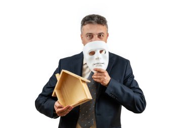 İş elbisesi giymiş orta yaşlı bir adam (emlakçı) elinde beyaz bir maske ve minyatür ahşap bir ev tutuyor.