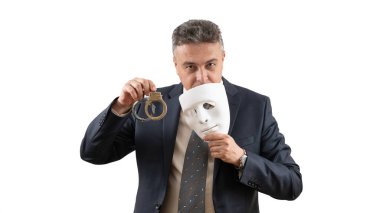 Takım elbiseli orta yaşlı bir adam elinde beyaz bir maske ve kelepçe tutuyor..