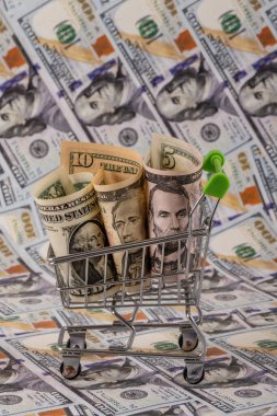 Minyatür bir alışveriş arabasında 1, 5 ve 10 dolarlık banknotlar ve arka planda 100 dolarlık banknotlar.