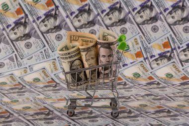 Minyatür bir alışveriş arabasında 1, 5 ve 10 dolarlık banknotlar ve arka planda 100 dolarlık banknotlar.