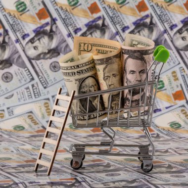 Minyatür bir alışveriş arabasında 1, 5 ve 10 dolarlık banknotlar ve arka planda 100 dolarlık banknotlar.
