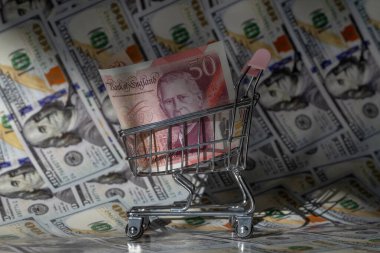 Minyatür bir alışveriş arabasında 50 İngiliz sterlini. Arka planda 100 dolar banknot var.