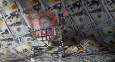 Minyatür bir alışveriş arabasında 50 İngiliz sterlini. Arka planda 100 dolar banknot var.