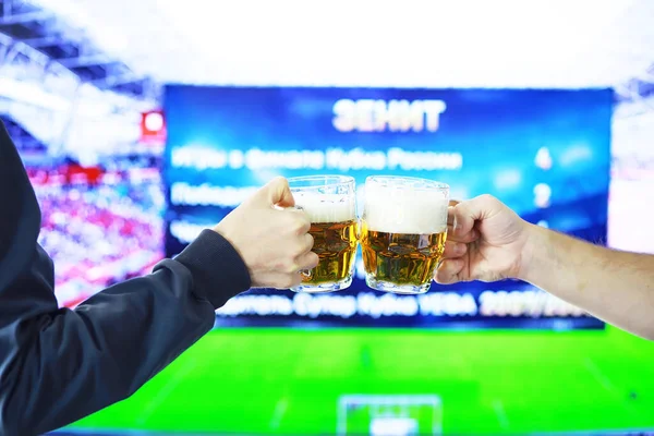 Hands horizontal holding lager beer glass and clinking on background of football game. Sport fans cheer up. Friends leisure lifestyle concept.