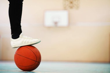 Beton zemindeki basketbol topunda bir insan ayağı dinleniyor. Tahta zeminde bir basket topu ve spor ayakkabısının fotoğrafı.