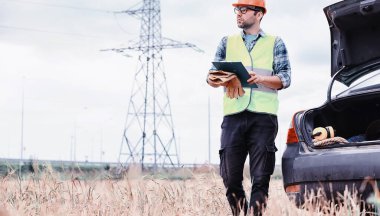 Kasklı ve üniformalı bir adam, sahada bir elektrikçi. Profesyonel elektrik mühendisi elektrik hatlarını inceliyor..