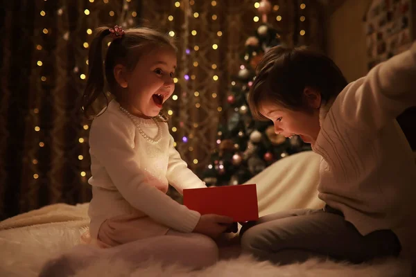 Family on Christmas eve at fireplace. Kids opening Xmas presents. Children under Christmas tree with gift boxes. Decorated living room with traditional fire place. Cozy warm winter evening home.