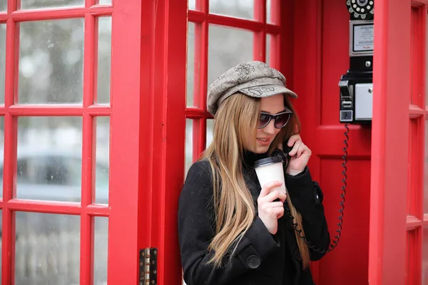 Beautiful young girl in a phone booth. The girl is talking on the phone from the payphone. English telephone booth in the street and a woman talking on the phone