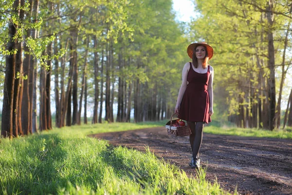 A girl in a hat on a walk in the park. A girl with a basket walks in the spring. Girl is walking along the road at sunset
