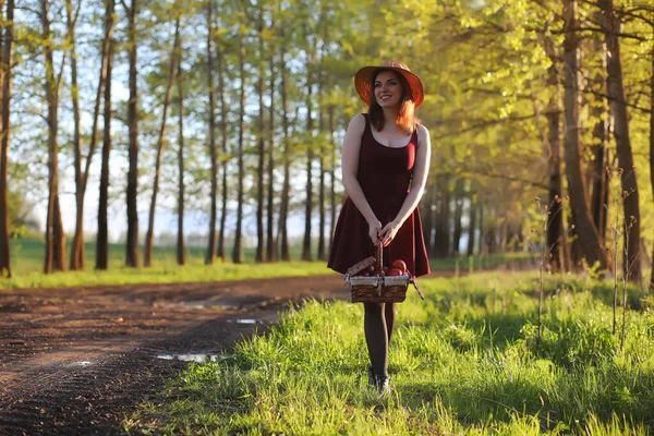 A girl in a hat on a walk in the park. A girl with a basket walks in the spring. Girl is walking along the road at sunset