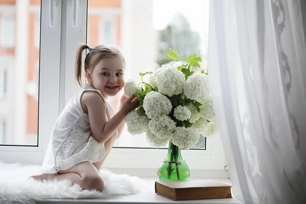 一个小女孩正坐在窗台上 窗边花瓶里的一束花和一个嗅着花的女孩 一个穿着白衣 拿着一束白花的小公主 — 图库照片