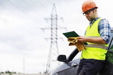 Kasklı ve üniformalı bir adam, sahada bir elektrikçi. Profesyonel elektrik mühendisi elektrik hatlarını inceliyor..