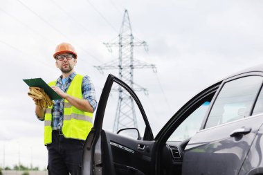 Kasklı ve üniformalı bir adam, sahada bir elektrikçi. Profesyonel elektrik mühendisi elektrik hatlarını inceliyor..