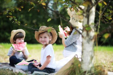 Parkta piknik yapan mutlu bir aile, bahçedeki çimlerde oturan ve dışarıda karpuz yiyen çocukları olan bir aile.