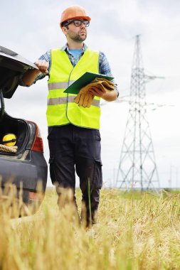 Kasklı ve üniformalı bir adam, sahada bir elektrikçi. Profesyonel elektrik mühendisi elektrik hatlarını inceliyor..