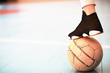 Beton zeminde bir insan ayağı dinleniyor. Ahşap zeminde bir futbol topu ve spor ayakkabısının fotoğrafı.