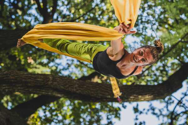 Aerial silk artist performing movements on ropes hanging from trees on a sunny day in Tivoli park, Ljubljana.