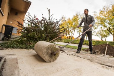 Bir adam eski beton silindiri kullanarak bir evin önündeki kumla kaplı zemini sertleştiriyor..