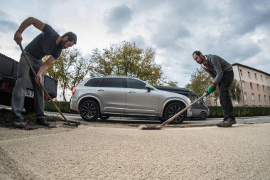 İki adam yerleşim yerinin önündeki kumu düzleştirmek ve düzleştirmek için tırmık ve kürek kullanıyor. Ev inşaat işçileri, bir araba geçiyor..