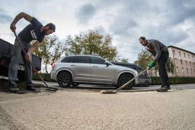 İki adam yerleşim yerinin önündeki kumu düzleştirmek ve düzleştirmek için tırmık ve kürek kullanıyor. Ev inşaat işçileri, bir araba geçiyor..