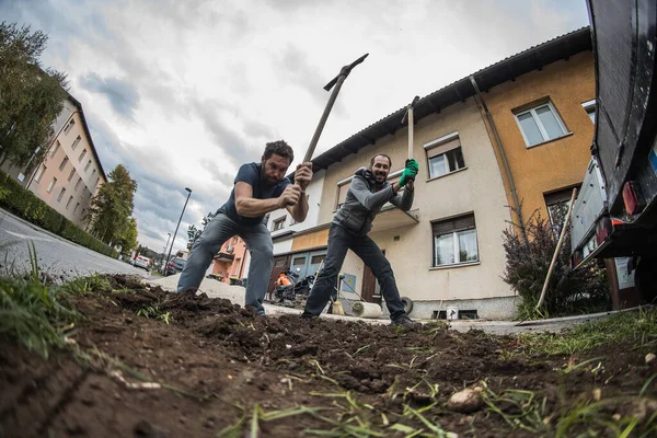 Two men are using a hack to soften the ground with a plan to dig it out in front of a residential house and cover the place with sand. Home construction.