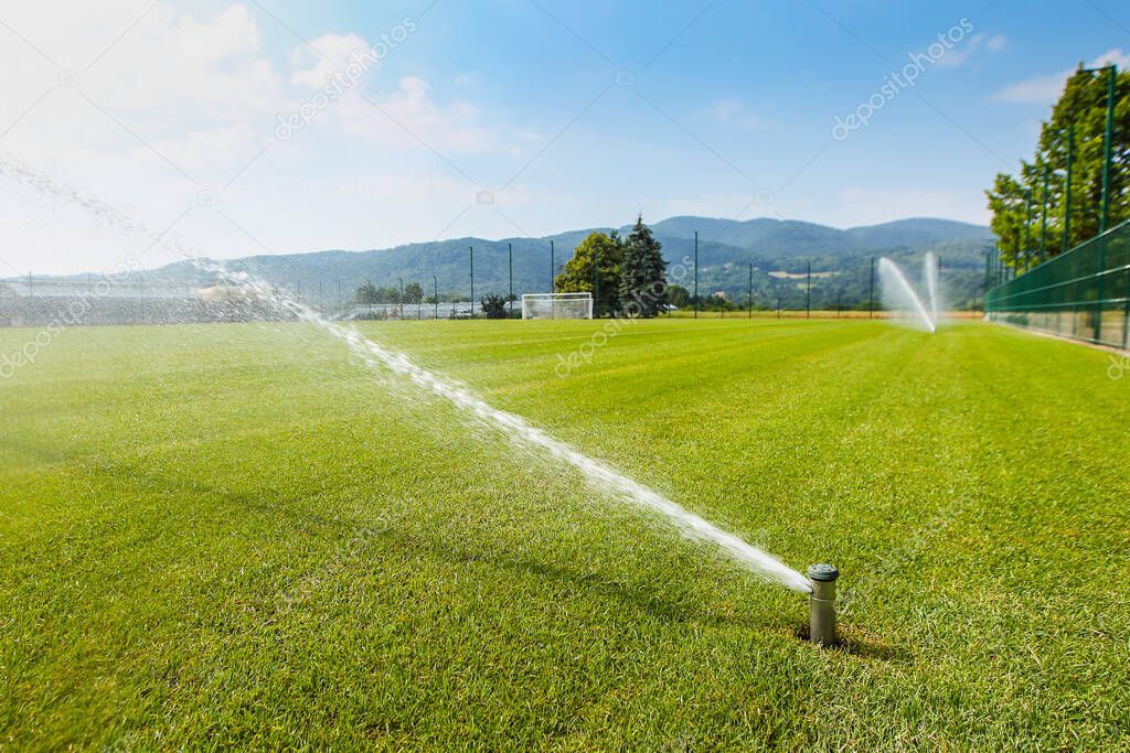 A sprinkler is watering a green football field, which is surrounded by a green fence. Hills in the background.
