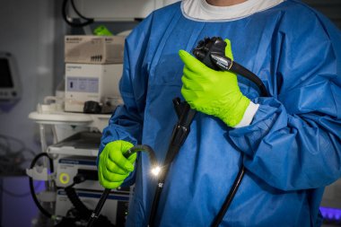 Yeşil eldivenli mavi elbiseli doktor elinde bir endoskop tutuyor. Endoskopun ışığı açık, arka planda tıbbi gereçler ve aletler var..