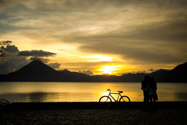 Panoramic view of Lake Atitlan in Guatemala with a couple holding each other in the evening with rich colors of sunset, volcano San Pedro and an old mountain bicycle in the foreground, resting on the stone wall.