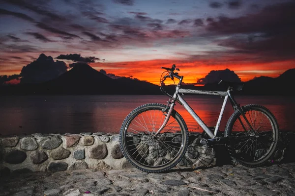 Panoramic view of Lake Atitlan in Guatemala in the evening with rich colors of sunset, volcano San Pedro and an old gray mountain bicycle in the foreground, resting on the stone wall.