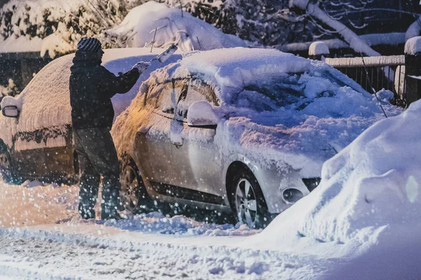 A person is cleaning snow and frost a passenger car on a street with the help of a brush in the morning getting ready for the morning commute. Cleaning the car from snow for a commute.