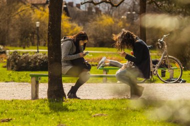 Güneşli bir günde parktaki bankta telefon tutan iki genç kadın. Güzel bir çevrede hüzünlü modern asosyal yaşam tarzı kavramı.