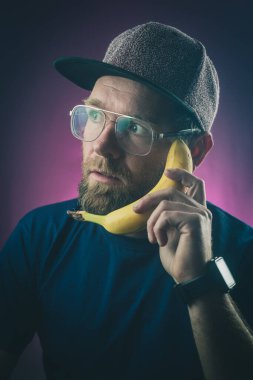 Hippi görünümlü, gözlüklü ve şapkalı genç bir erkek, mor veya pembe arka planda muz kullanıyor. Muzla iletişim kurma kavramı.