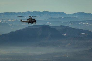 Askeri kurtarma helikopteri, tepe ve dağ sırtlarıyla dolu muhteşem bir arka plan önünde, gökyüzünün yan kapısı açık bir şekilde havada asılı duruyor..