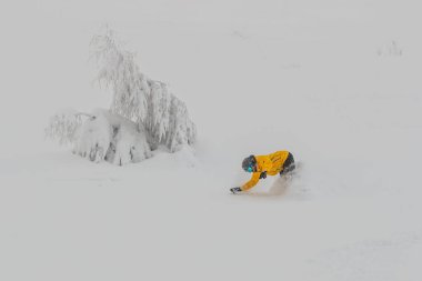 Toz kar içinde bir snowboardcu viraja girmek üzere. Siyah pantolonlu, sarı elbiseli ve bulutlu bir günde tozun içine düşen turuncu panelli bir pansiyoner..