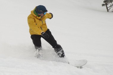 Kar içinde bir snowboardcu yokuş aşağı atlıyor. Siyah pantolonlu, sarı elbiseli, turuncu sörf tahtası olan bulutlu bir günde karda atlayan ve zıplayan biri..