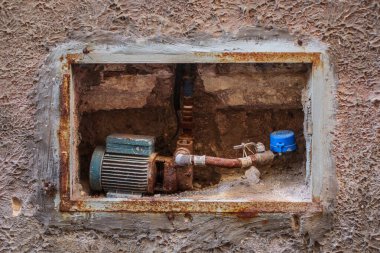 Evin duvarına monte edilmiş eski bir elektrikli su pompası. Eski paslı su pompası görünür boruları, vanası ve tezgahı var. Evlerdeki eski boru sistemi kavramı.