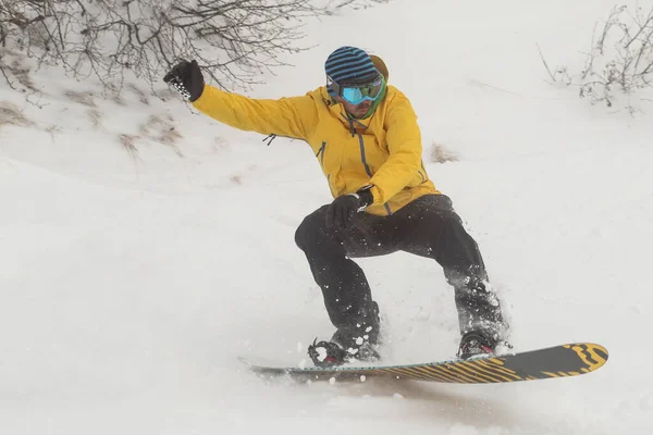 ジャンプの間に下り坂に行く雪の中でスノーボーダー 黒いズボンとオレンジボードに乗って雪の上に曇りの日にジャンプして黄色の服でボーダー — ストック写真