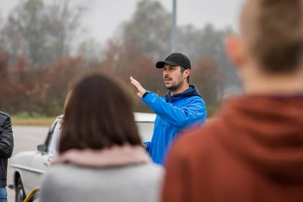 A white male man is explaining directions or route to other listeners on a parking lot on an early foggy autumn morning.