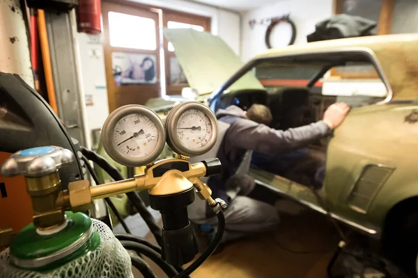 Welding tank pressure gauges are seen in the foreground while a  man is inspecting an old vintage car with an attempt to restore it.