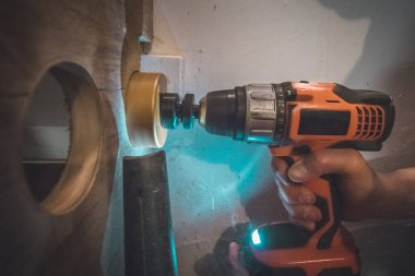 Aküye dayanıklı matkap kullanan bir adam tahta bir kalasa delik açmak için testere ucu kullanmış. Elektrikli süpürge fazla tozu emmek için görünür..