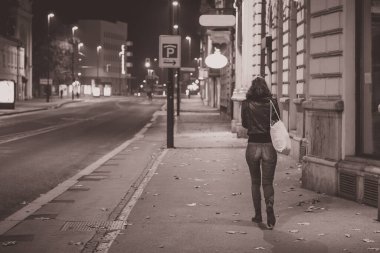 Kotlu ve ceketli yalnız bir kadın geceleri boş bir caddede kameradan uzaklaşıyor.
